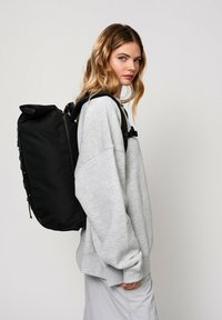 Zaino nero con superficie testurizzata, chiusura a rotolo e spallacci regolabili. Indossato sopra una felpa oversize grigia chiara.
