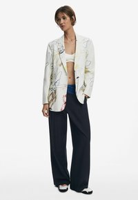 Blazer blanc avec des motifs floraux colorés, associé à un top court blanc. Pantalons noirs amples et chaussures noir et blanc.