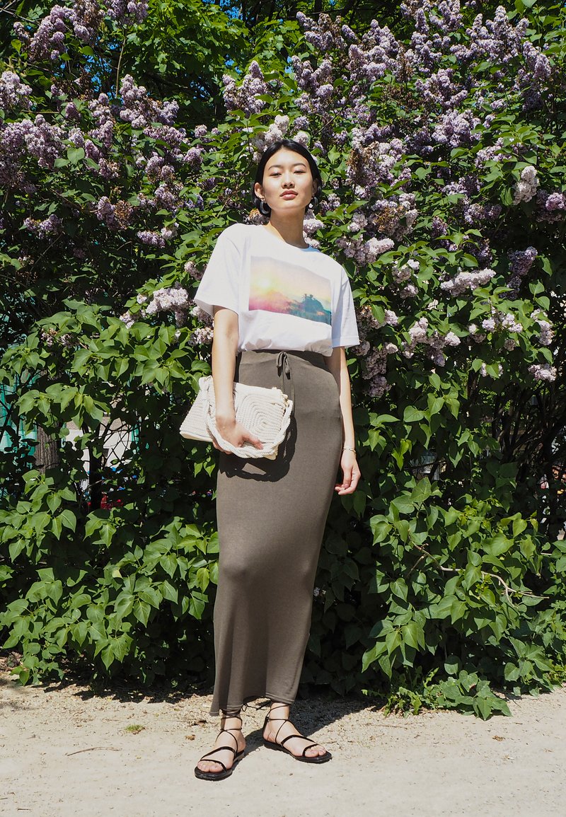 Vit t-shirt med tryck av en färgglad landskap, olivgrön bodycon kjol, texturerad beige väska och svarta remsandaler mot en bakgrund av lila blommor.