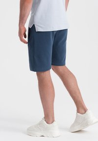 Modré bavlněné šortky s volným střihem, mají boční kapsy a hladkou texturu. Doplňuje je bílá robustní tenisová obuv.