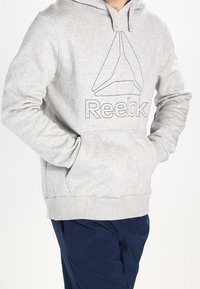 Šedá mikina s kapucí vyrobená z měkké bavlněné směsi, s předními klokaními kapsami a potiskem loga Reebok. Součástí je nastavovací šňůrka na kapuci.