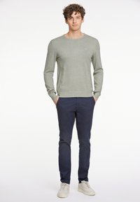 Maglione grigio chiaro a maglia con colletto rotondo, abbinato a pantaloni slim-fit blu scuro e sneakers bianche, in piedi contro uno sfondo neutro.