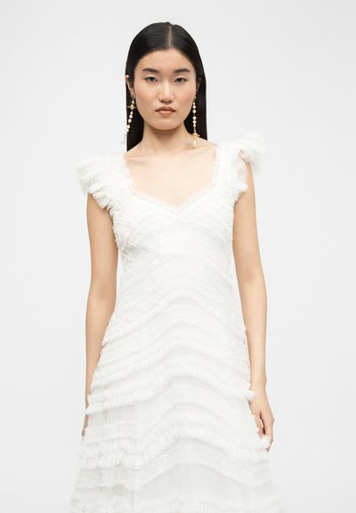 Γυναίκα με μακριά μαύρα μαλλιά που φοράει λευκό αμάνικο φόρεμα με βολάν και μακριά σκουλαρίκια με μαργαριτάρια, μπροστά σε απλό φόντο.