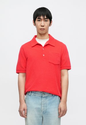 Rød kortermet poloshirt med krage og tre knapper, med en brystlomme. Brukt med lyseblå denimjeans.