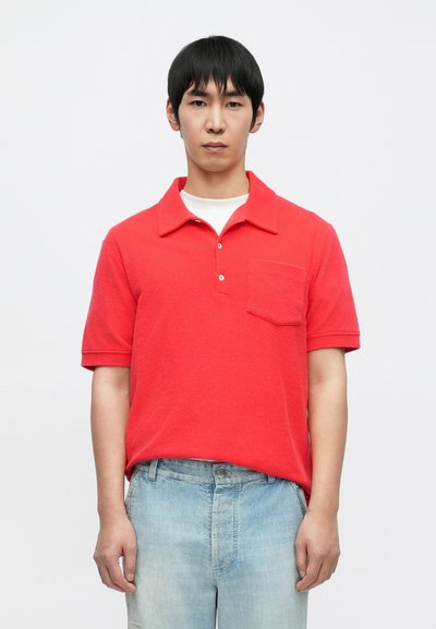 Polo rouge à manches courtes avec col et trois boutons, doté d'une poche poitrine. Porté avec un jean denim bleu clair.