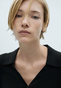 Close-up van een persoon met kort lichtblond haar, in een zwart overhemd met kraag. De huid heeft een gladde textuur, met neutrale make-up.