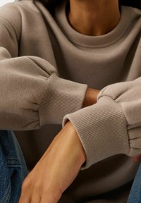 Persona que lleva un sudadera beige con puños acampanados y jeans azules, descansando el brazo sobre su regazo.