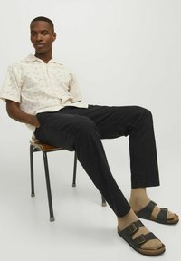 Camisa perforada de manga corta color crema sobre pantalones negros de pierna recta, combinada con sandalias marrones y negras de doble hebilla, usadas con calcetines beiges.