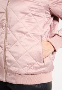 Chaqueta bomber acolchada de color rosa claro con cremallera dorada, puños de canalé y bolsillos laterales, hecha de una tela suave y brillante.