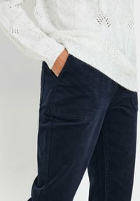 Manos que llevan un suéter blanco de punto metido en pantalones de pana azul marino, con la mano en el bolsillo, contra un fondo claro.