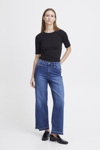 Schwarzes, eng anliegendes Oberteil mit kurzen Ärmeln, kombiniert mit weiten, hellblauen Jeans mit rohem Saum. Modell steht in schwarzen Absatzschuhen vor weißem Hintergrund.