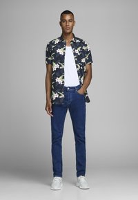 Camisa de botones de manga corta con estampado floral oscuro sobre una camiseta blanca; jeans de mezclilla azul y zapatillas blancas; de pie en un fondo neutral.