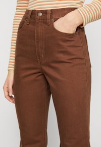 Pantalones de denim marrón con una cintura alta, diseño de pierna recta, costuras visibles y bolsillos frontales. La tela parece suave con una ligera textura.