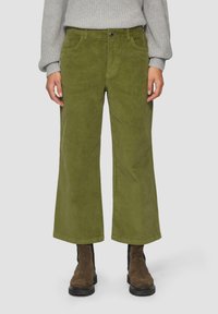 Olivgrüne Cordhose mit weitem Bein und sichtbaren Taschen, mit einem Frontknopfverschluss, kombiniert mit braunen Stiefeletten.