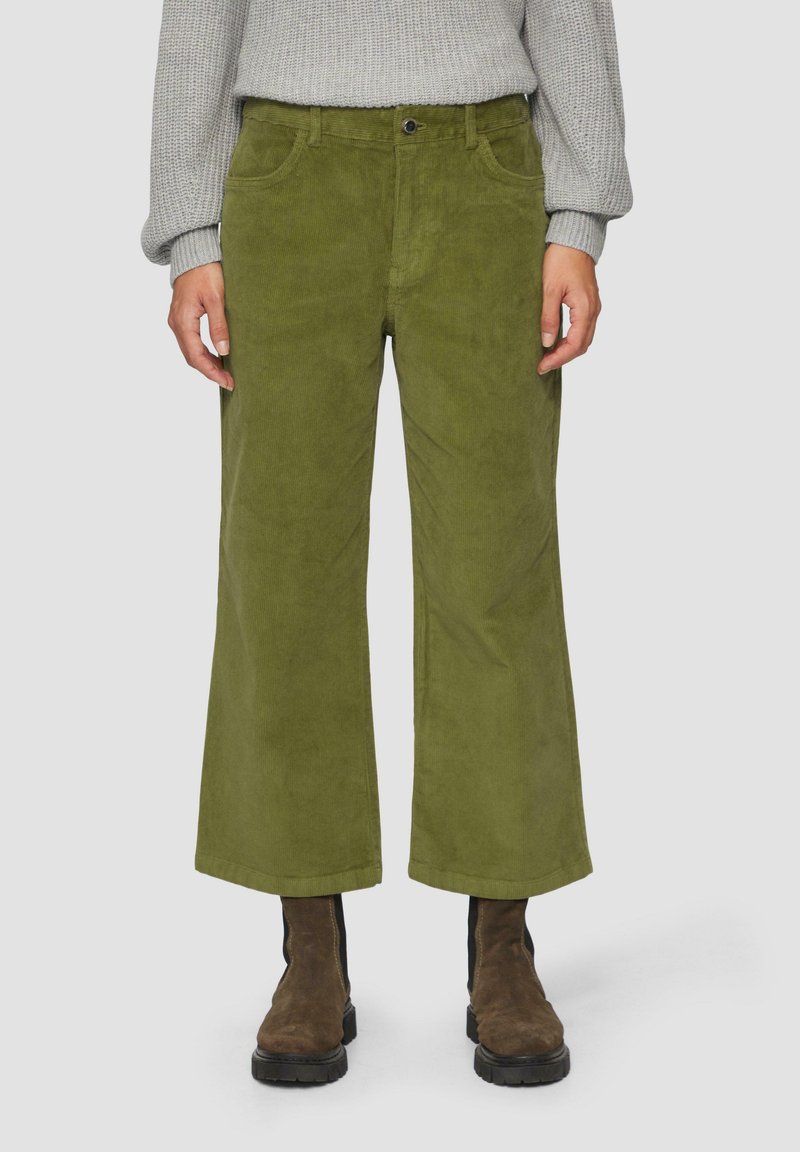 Olivgrüne Cordhose mit weitem Bein und sichtbaren Taschen, mit einem Frontknopfverschluss, kombiniert mit braunen Stiefeletten.
