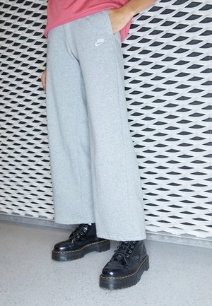Personne portant un pantalon de jogging Nike gris à jambes larges et des bottes à plateforme noires, debout contre un mur blanc à motifs.