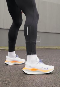 Laufschuhe mit weißem Netz-Obermaterial, orangefarbener Zwischensohle und lila Schnürsenkeln. Schwarze enganliegende Leggings mit reflektierenden Akzenten und Reißverschlussdetails an den Knöcheln.