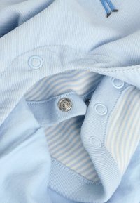 Hellblauer Stoff mit gerippter Textur, ausgestattet mit Druckknöpfen und gestreifter Innenauskleidung. Beinhaltet ein kleines gesticktes Design im Stoff.