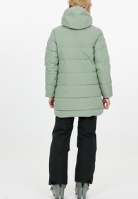 Vadderad kappa i ljusgrön, med huva, elastiska ärmslut och en medellång design. Bärs tillsammans med svarta byxor och grå stövlar.