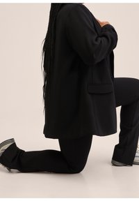 Personne aux cheveux longs et tressés à genoux, portant un blazer noir, un pantalon noir et des baskets, sur fond beige.