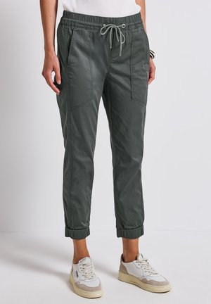 Dark green elastic waistband pants with a drawstring, side pockets, and elastic cuffs, made from a lightweight material. Worn with white sneakers.