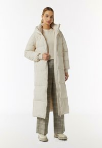 Puffermantel in hellem Beige mit Kapuze, mit einem Reißverschluss vorne, gestepptem Textur und gerippten Bündchen. Kombiniert mit karierten Hosen und crèmefarbenen Schuhen.