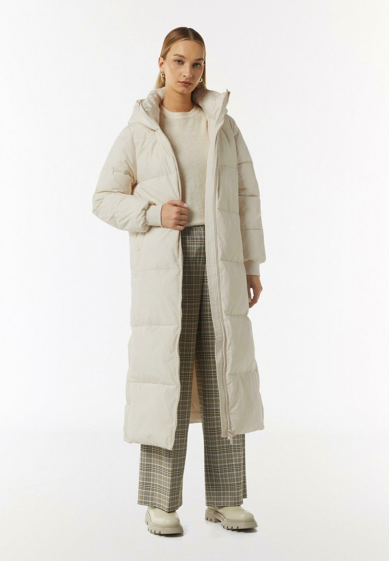 Puffermantel in hellem Beige mit Kapuze, mit einem Reißverschluss vorne, gestepptem Textur und gerippten Bündchen. Kombiniert mit karierten Hosen und crèmefarbenen Schuhen.