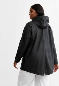 Svart huvad regnjacka med en lös passform, tillverkad av slätt, vattentätt material. Har elastiska ärmslut och en ventil på baksidan för ökad andningsförmåga.