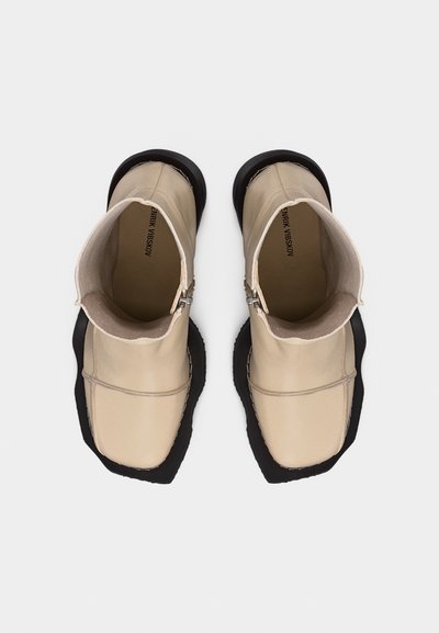 Μπεζ δερμάτινες μποτάκια αστραγάλου με τετράγωνα μύτα, μαύρες ανθεκτικές σόλες και φερμουάρ στο πλάι, εμφανιζόμενα από πάνω σε λευκό φόντο.