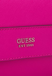 Jasnoróżowy materiał syntetyczny o teksturowym wykończeniu. Złote logo "GUESS EST 1981". Płaski design z widocznymi detalami ściegów.