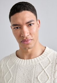 Cremfarbener Kabelstrickpullover mit ribbeligem Ausschnitt und strukturiertem Muster. Nahaufnahme des Gesichts eines männlichen Modells vor einem neutralen Hintergrund.