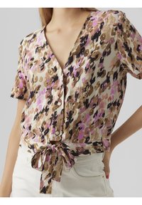 Donna che indossa una blusa a maniche corte con bottoni, con un motivo floreale astratto nei colori rosa, marrone e nero, annodata in un fiocco in vita, abbinata a pantaloni bianchi.