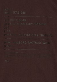 Maroon t-shirt med svart tryckt text; detaljer inkluderar "HALO," "OFF DUTY GEAR," och "PHYSICAL EDUCATION & TRAINING." Slät textur.
