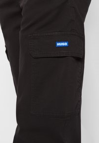 Black cargo pants with a side pocket, featuring a blue "HUGO" logo. The material is smooth with a sturdy texture and tailored fit.