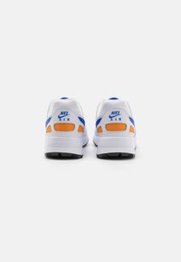Bílé tenisky Nike Air s modrými a oranžovými akcenty; mají texturovaný svršek, polstrovaný límec a gumovou podrážku pro lepší přilnavost.
