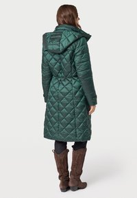 Dlouhá zelená prošívaná puffer bunda s kapucí, páskem v pase a diamantovým vzorem; doplněná hnědými kovbojskými botami. Hladká, lesklá textura.
