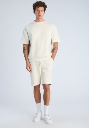 Uomo con capelli castani ricci in piedi con t-shirt e pantaloncini color crema, calzini bianchi e sneakers, mani in tasca, tatuaggi su entrambi gli avambracci.