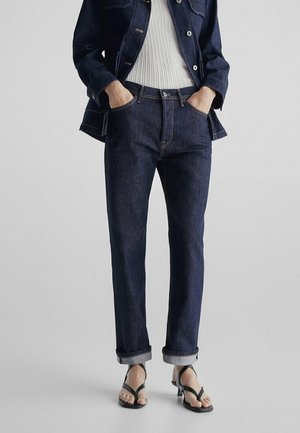 Personne portant un jean en denim bleu foncé avec des revers, une veste en denim assortie, un haut blanc côtelé et des sandales à talons noires à lanières, les mains dans les poches.