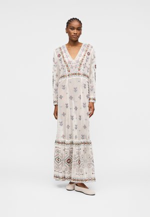 Femme portant une longue robe blanche avec des broderies florales colorées et des motifs, debout avec une expression neutre et des chaussures plates blanches.
