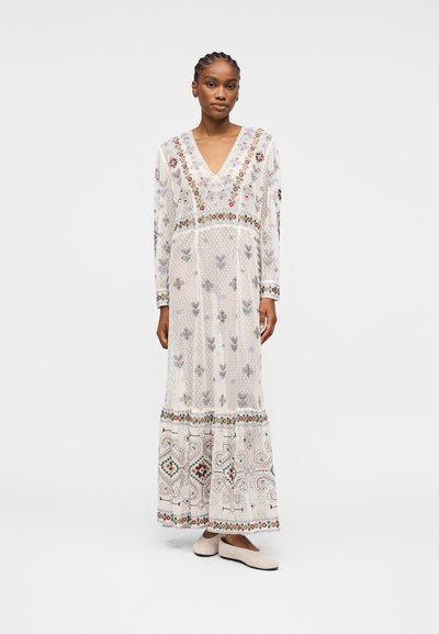 Femme portant une longue robe blanche avec des broderies florales colorées et des motifs, debout avec une expression neutre et des chaussures plates blanches.