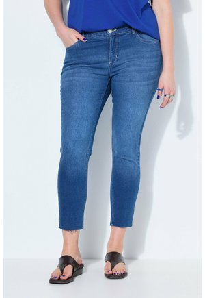 Kvinde iført blå forkortede skinny jeans med rå kanter, en blå top og sorte sandaler med åben tå, stående med den ene hånd i lommen.