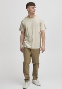 Beige bomulds T-shirt med rund halsudskæring og korte ærmer, med et lille sort logo på venstre bryst. Sammen med lysebrune bukser.