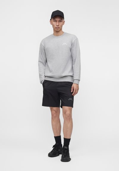 Άνδρας που φορά γκρι φούτερ, μαύρες βερμούδες, μαύρες κάλτσες, μαύρα αθλητικά παπούτσια και μαύρο καπέλο μπέιζμπολ, στέκεται μπροστά σε απλό φόντο.