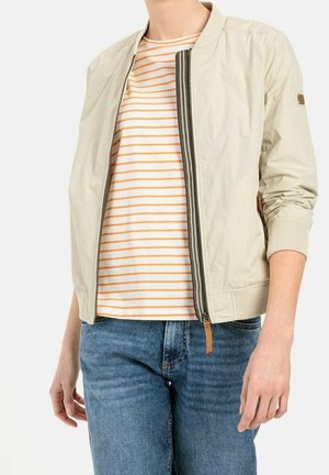 Personne portant une veste beige zippée sur une chemise blanche à rayures orange et un jean bleu, vue du cou aux hanches, sur un fond uni.