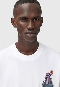 Hvid bomulds T-shirt med rund halsudskæring, der har et grafisk design af en lilla næve, der holder en rose, omgivet af tornede detaljer.
