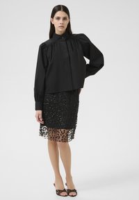 Blouse noire courte avec des manches bouffantes et un col, associée à une jupe noire ornée de sequins avec un ourlet transparent.