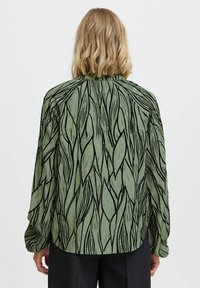 Blouse verte avec un motif de feuilles noires. Manches longues et coupe décontractée, avec un tissu texturé. Sans boutons ni ornements visibles.