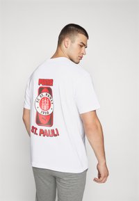 Balts T-krekls ar uzdrošinošu sarkano grafiku FC St. Pauli logo aizmugurē, zem kura ir teksts "PUMA" un "ST. PAULI".