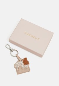 Coccinelle nøkkelring i beige skinn, husformet, har teksturert mønster og brune detaljer. Festet til en sølvfarget nøkkelring. I eske.
