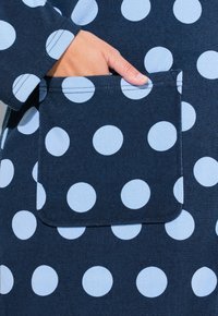 Tessuto blu navy con pois azzurri. Presenta una tasca frontale perfettamente integrata nel design, con dettagli di cucitura visibili.